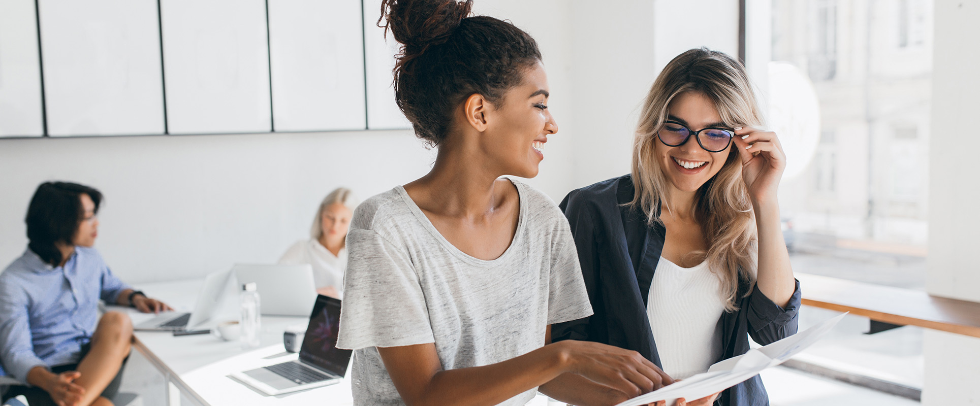 Young female executive explaines new strategy to blonde employee in glasses and smiling. Indoor portrait of multicultural collective working on project in office and using laptop..